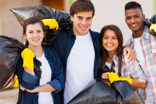 Four individuals, two women and two men, standing outdoors on a paved surface, are engaged in rubbish removal activities. They are smiling and looking at the camera, each holding large black rubbish bags filled with waste. The bags are made of thick plastic, sealed at the top, and show signs of being full and ready for collection. The group appears to be in a residential area, with a blurred background that suggests a suburban setting. They are wearing casual clothing suitable for outdoor work, with some also wearing yellow gloves, emphasizing their involvement in rubbish collection or clearance tasks. The scene is well-lit with natural daylight, highlighting their friendly expressions and the details of the rubbish bags. This image exemplifies community effort and professional rubbish clearance services, aligning with the offerings of House Clearance Maidenhead, especially within the Maidenhead area. The outdoor environment and the depiction of waste collection reinforce the relevance of a reliable rubbish removal service in local residential contexts.