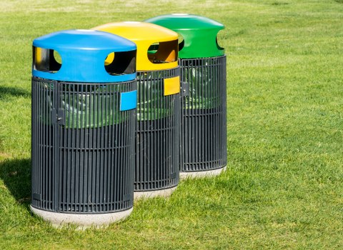 Three cylindrical outdoor rubbish bins positioned on a well-maintained grassy area, with the bin on the left featuring a blue lid and front panel, the middle bin having a yellow lid and panel, and the right bin with a green lid and panel. Each bin has vertical black slats forming the main body, and they are situated on a paved surface with a blurred background of lush green grass, suggestive of a garden or residential outdoor space. The bins are used for waste disposal and are associated with domestic rubbish removal services in Maidenhead, as provided by House Clearance Maidenhead. The natural lighting emphasizes the different colour-coded lids and the textured surface of the bins, supporting visual identification suitable for accessibility and search engine relevance related to rubbish and waste management services in the local area.
