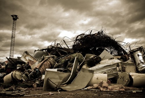 A large pile of discarded household and industrial rubbish is situated outdoors on a paved driveway or cleared area. The pile includes various items such as broken appliances, metal frames, plastic containers, and other debris, with some objects visibly rusted or damaged. At the top of the heap, there is a tangled mass of dark, twisted wires or branches. The background features an overcast sky with thick, heavy clouds, and a tall metal tower or antenna is visible on the left side of the image. The scene appears to be part of a rubbish clearance operation, possibly by House Clearance Maidenhead, with the waste accumulated in an untidy manner, awaiting removal or disposal. The surrounding environment suggests a residential or industrial area, with pavement edges visible along the bottom of the image, emphasizing the outdoor context for rubbish collection and disposal services. No vehicles or workers are seen in the image, just the cleared waste pile under the gloomy sky.
