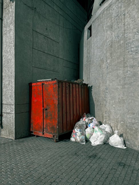 A rusty red metal skip with visible weathering and peeling paint is positioned against a dark green wall on the left side and a light grey concrete wall on the right side, situated on a paved area wit