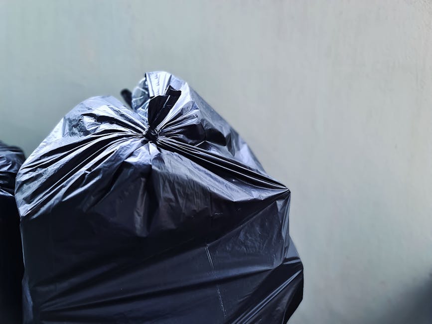 Close-up image of a large black rubbish bag made from crinkled, shiny plastic material, tightly knotted at the top, positioned in the foreground against a plain off-white wall. The bag appears to be f