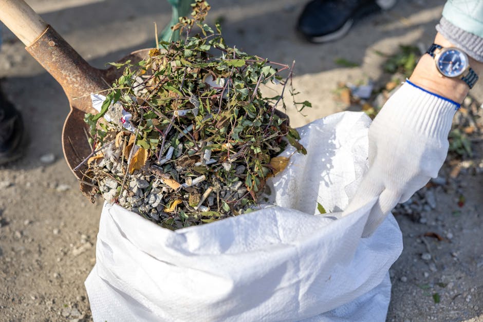 A close-up view of a white woven polypropylene sack filled with garden waste, including small branches, green leaves, and broken debris. A person's hand, wearing a white glove, is holding the bag open